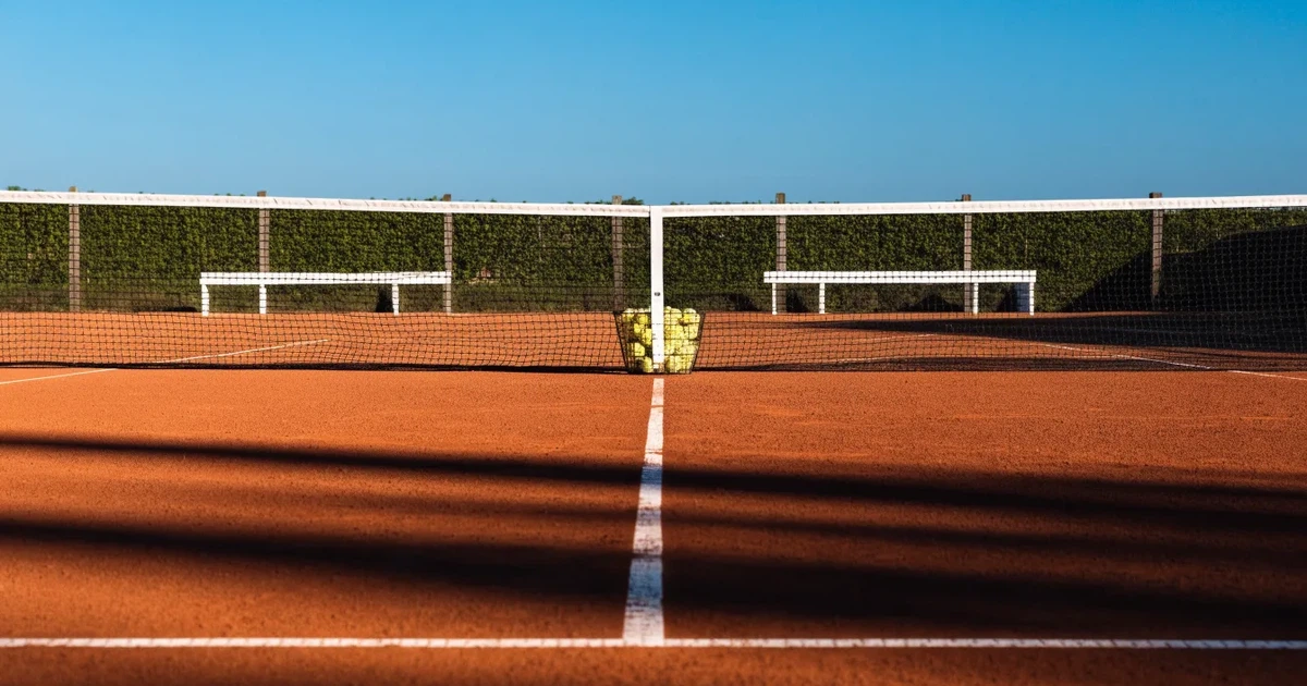 Ein Sandplatz mit frisch gezogenen Linien in der Nachmittagssonne, Netz und Ballkorb am Rand, keine Menschen.